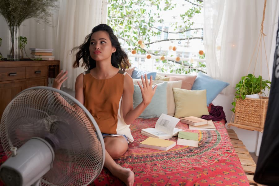 young woman surrounded by books on a bed trying to cool off with a fan